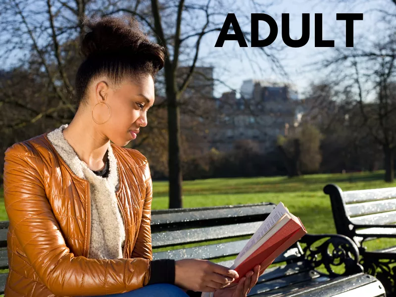 African-American woman reading a book on a bench in a park. Woman is wearing hoop earrings, a brown coat, and beige scarf. 