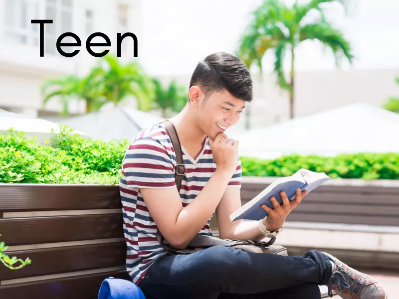 Young man sitting outside at a school. He is reading a book and sitting on a bench, wearing dark jeans and a red, white, and blue striped shirt